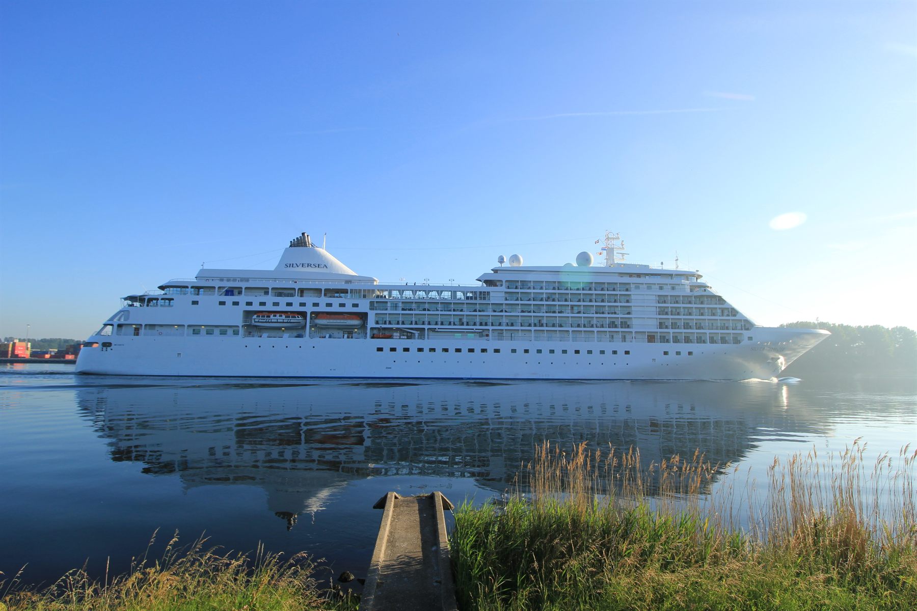 A Silversea cruise ship reflected in calm water beside a grassy shore
